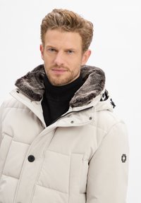 Man met lichtbruin haar en baard, gekleed in een beige gewatteerde jas met een met bont gevoerde capuchon en een zwarte coltrui, die recht vooruit kijkt.