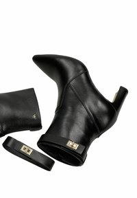 Černé kožené kotníkové boty s špičatou špičkou a zakřiveným podpatkem. Mají malý kovový akcent a odnímatelný dekorativní pásek.