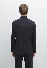 Mörkgrå skräddarsydd kavaj med slät yta, diskret rutmönster, jackslag med knapphål och knäppta manschetter, sedd bakifrån.