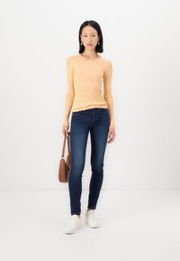 Light orange long-sleeve top with a scalloped neckline, paired with dark blue skinny jeans and white sneakers. Brown handbag included.