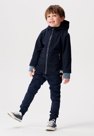 Hoodie navy avec fermeture éclair et poches latérales, associé à un jogger navy assorti. Le design des poignets gris et des baskets blanches complètent le look.
