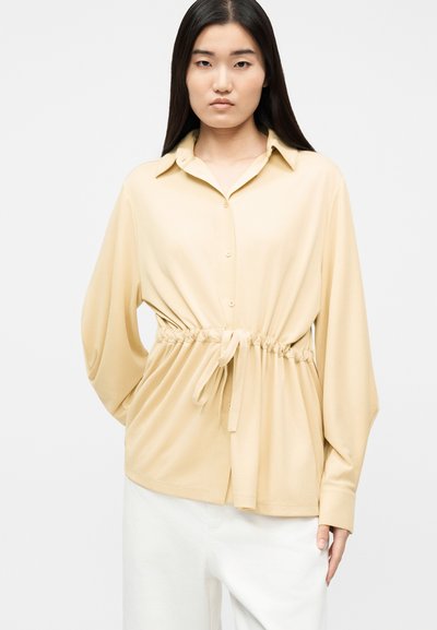 Γυναίκα με μακριά σκουρόχρωμα μαλλιά, φορώντας μπεζ πουκάμισο με κουμπιά και ζωνάκι στη μέση, καθώς και λευκό παντελόνι, στέκεται μπροστά σε απλό φόντο.