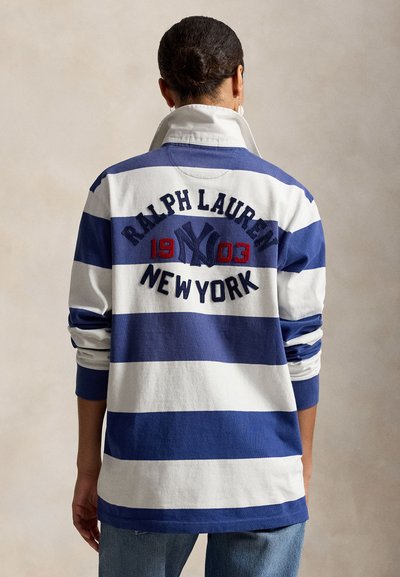 Personne aux cheveux courts portant un maillot de rugby à rayures bleues et blanches avec "Ralph Lauren New York 1903" brodé dans le dos.