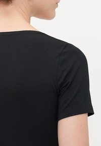 Nahaufnahme des oberen Rückens und der Schulter einer Person, die ein einfaches schwarzes T-Shirt mit kurzen Ärmeln vor einem weißen Hintergrund trägt.