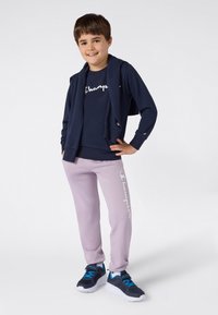 Leende pojke iklädd marinblå sweatshirt och lila sweatpants med varumärkeslogo, marinblå jacka draperad över axlarna och marinblå sneakers med blå snören.