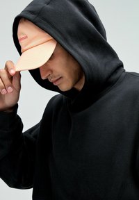 Zwarte sweatshirt met capuchon, met een aansluitend ontwerp. Een perzikkleurige pet met een logo wordt gedragen, waardoor het gezicht gedeeltelijk wordt verborgen.