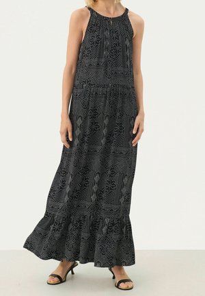 Femme portant une robe maxi noire sans manches avec des motifs floraux et géométriques blancs, associée à des sandales à talons noires.