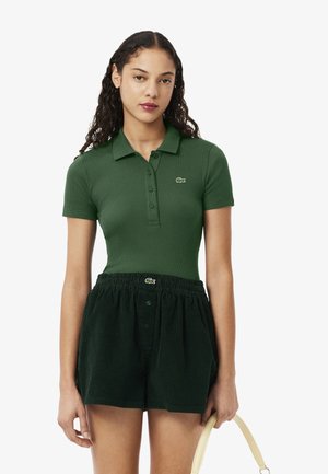Polo verde a costine con colletto e bottoni, abbinato a pantaloni corti in velluto a coste verde scuro con elastico in vita e dettaglio del logo.