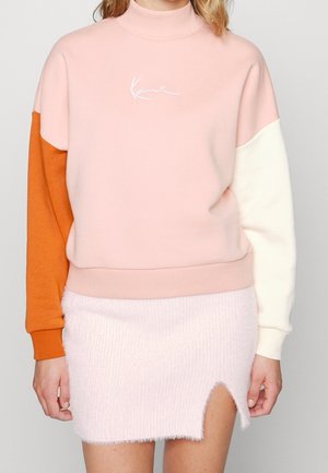 Vrouw draagt een roze sweatshirt met één oranje en één witte mouw, gecombineerd met een lichtroze zachte minirok met een voorsplit.
