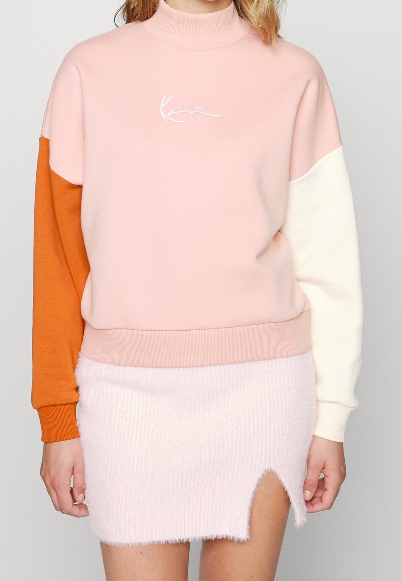 Femme portant un sweat-shirt rose avec une manche orange et une manche blanche, associé à une mini-jupe rose clair duveteuse avec une fente à l'avant.
