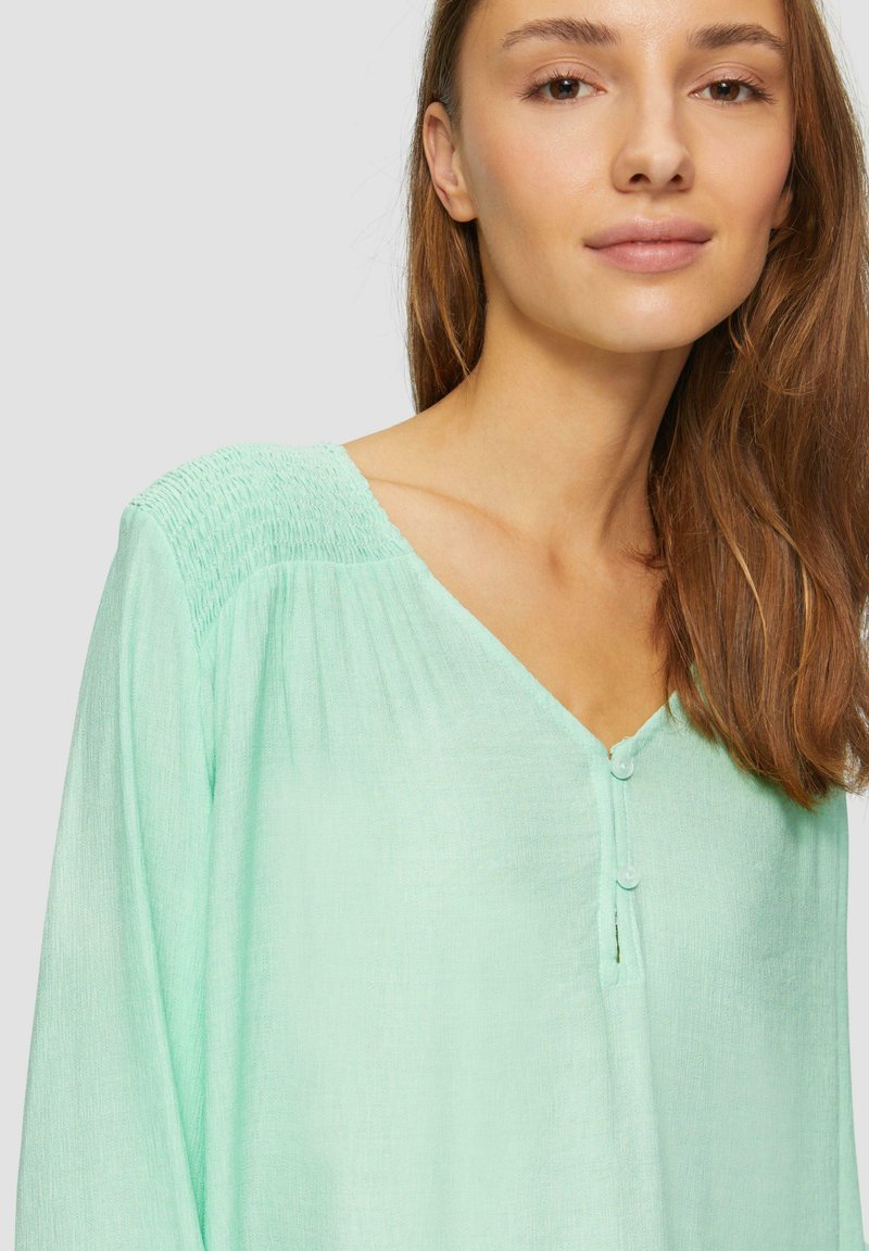 Mintgrünes Blusentop mit V-Ausschnitt, mit gerafften Schultern, langen Ärmeln und zwei kleinen Knöpfen auf der Vorderseite. Weicher, strukturierter Stoff.
