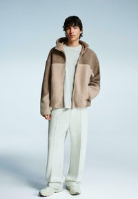 Jachetă din polar maro, cu fermoar și glugă, tricou deschis la culoare gri, pantaloni deschis la culoare gri și adidași albi; prezintă panouri și cusături contrastante.