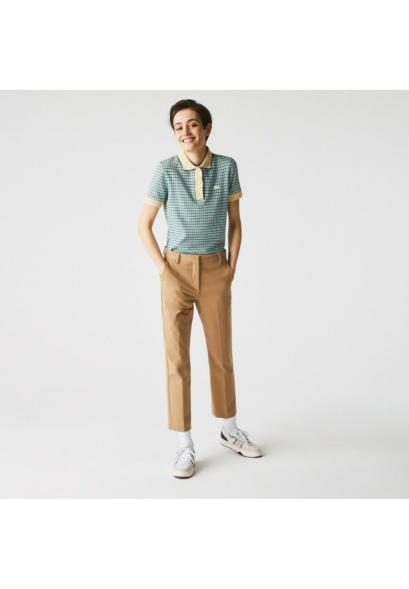 Polo shirt à carreaux verts avec un col crème, associé à un pantalon beige. Le modèle porte des baskets blanches avec des accents colorés.
