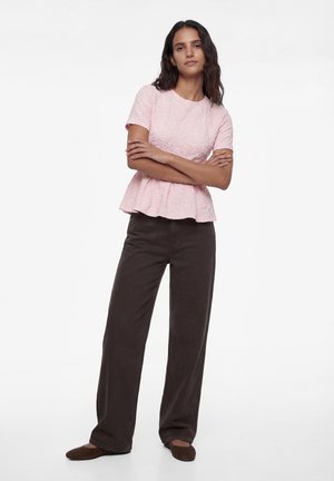 Kobieta z długimi ciemnymi włosami stoi z założonymi ramionami, ubrana w teksturowany różowy krótki top z krótkim rękawem, ciemne szerokie spodnie i brązowe baleriny.