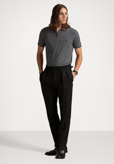 Polo gri cu guler cu fermoar, pantaloni plisati negri și pantofi negri slip-on, stând pe un fundal neutru.