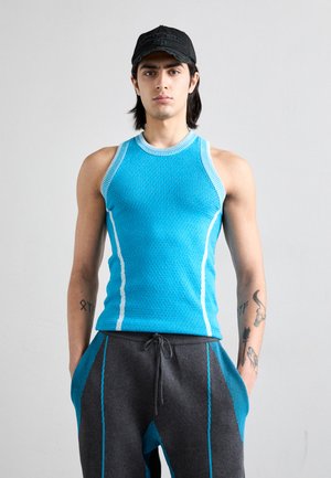 Blå tekstureret tanktop med hvide accenter, parret med grå sweatpants med blå sidelommer. Modellen står med hænderne i bukselommerne.