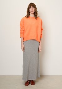 Svetlo oranžni udoben pulover iz mehkega materiala, v kombinaciji z dolgim sivim džersijevim krilom in rjavimi sandali s trakovi. Preprost, sproščen kroj.