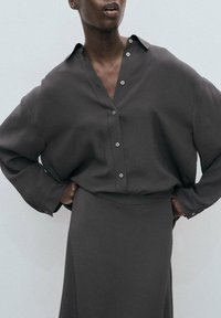 Camisa de botones de gran tamaño en gris oscuro con cuello, combinada con una falda a juego. Tejido suave, corte relajado y sutil detalle de botones.