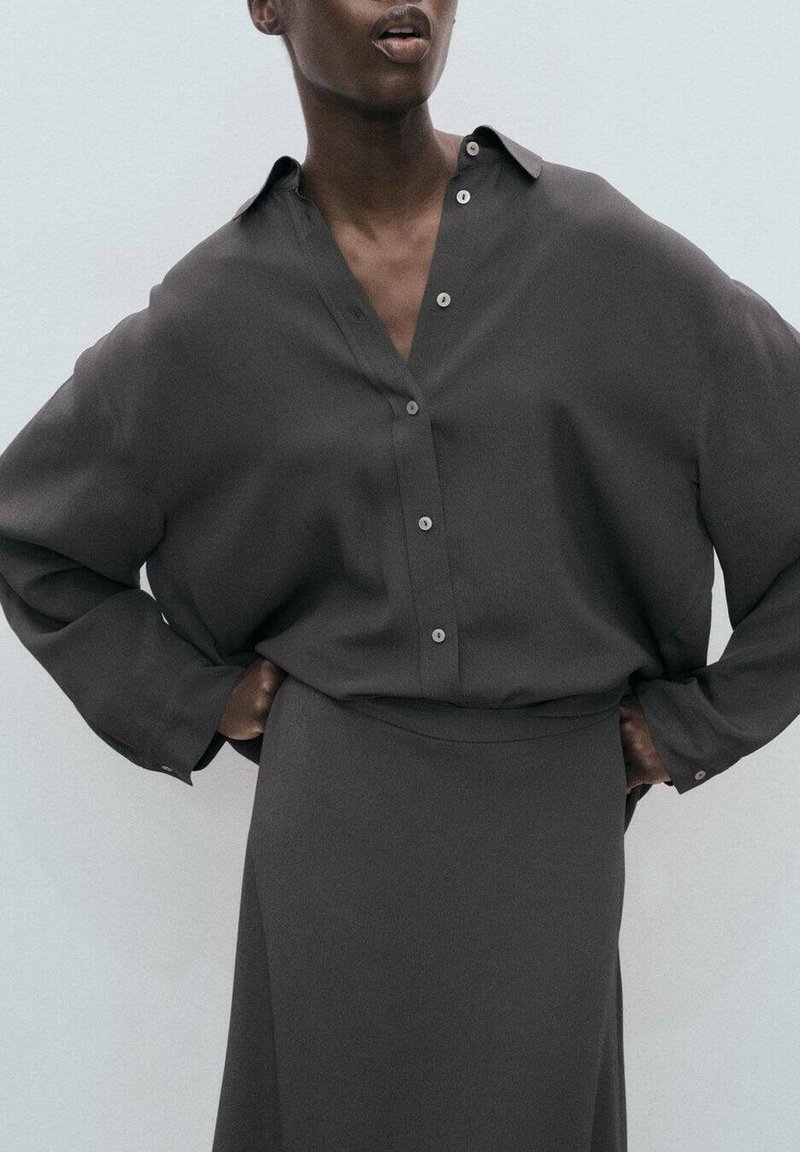 Donkergrijs oversized button-up shirt met een kraag, gecombineerd met een bijpassende rok. Soepele stof, ontspannen pasvorm en subtiele knoopdetails.