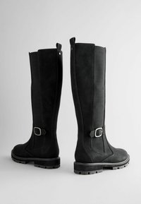 Black knee-high boots featuring a suede exterior, a strap detail with a silver buckle, and a textured side panel. Sturdy rubber sole.