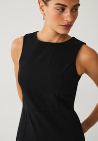 Sort ærmeløs kjole med høj hals, glat tekstur, skræddersyet pasform og diskrete sømme, som fremhæver en strømlinet silhuet.