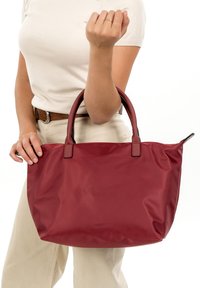 Raudonas nailoninis pirkinių krepšys su odinėmis rankenomis, turintis lygiu paviršiumi ir minimalistiniu dizainu. Krepšys laikomas viena ranka, dalinai atvertas.