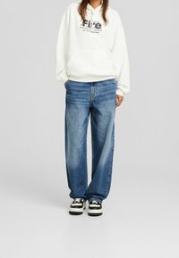 Sudadera blanca con estampado gráfico; jeans de pierna ancha azul; zapatillas negras y blancas. Estilo casual, ajuste relajado con una textura suave.