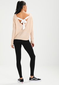 Un pull en tricot rose clair avec une découpe profonde dans le dos et un détail en nœud blanc, associé à un legging noir et des baskets noires.