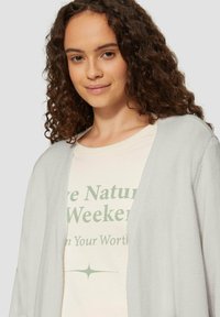 Jasnoszary kardigan z otwartym przodem, miękka tekstura. Noszony na beżowej koszulce z zielonym napisem i grafiką gwiazdy.