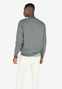 Sudadera gris de cuello redondo con puños y dobladillo acanalados, usada sobre una camisa de color verde claro, combinada con pantalones blancos, vista desde la parte de atrás.