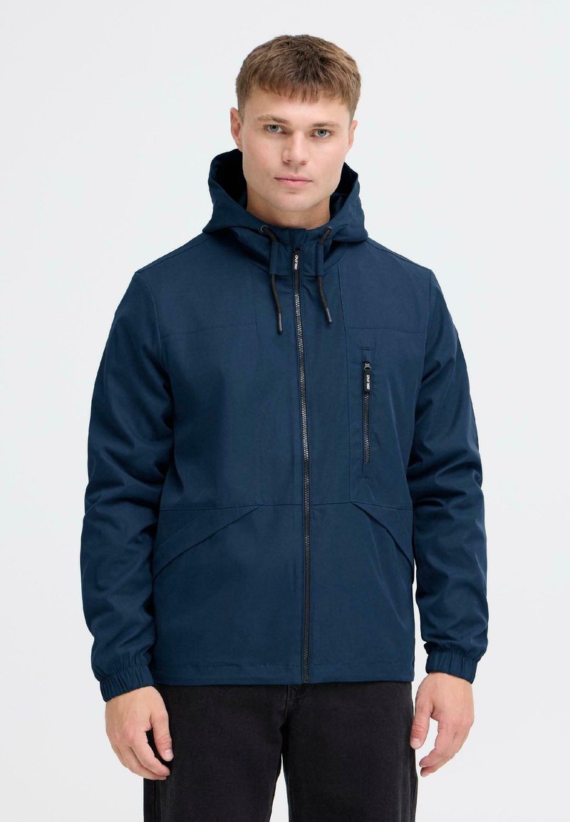 Mornarsko modra zip-up jakna s kapuco, ki ima žep na prsih in elastične manšete. Gladka tekstura z minimalističnim dizajnom in šivnimi detajli.