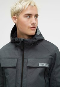 Szara wodoodporna kurtka z zamkiem z przodu, dwoma kieszeniami na piersi, regulowanym kapturem i detalem z logo po lewej stronie. Matowe wykończenie tkaniny.