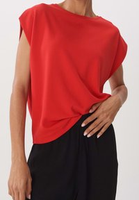 Rotes Kurzarmoberteil aus einem weichen, fließenden Stoff mit lockerer Passform und rundem Ausschnitt, kombiniert mit schwarzen Hosen, Hand in der Taille.