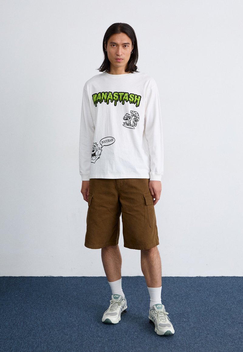 Chemise blanche à manches longues avec un graphique vert "WANASTASH", designs de dessins animés noirs. Shorts cargo marron. Baskets grises avec des accents blancs et chaussettes mi-mollets.