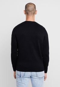 Pull en maille noir à col rond, avec des poignets et un ourlet côtelés, présentant une fente latérale et un tissu texturé. À porter avec un jean bleu clair.