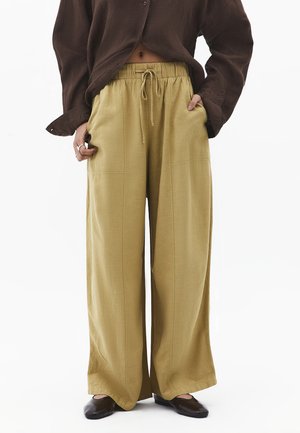 Osoba nosząca luźne, szerokie musztardowe spodnie z elastycznym pasem i sznurkiem, w połączeniu z ciemnobrązowymi butami i krótkim, ciemnobrązowym topem.