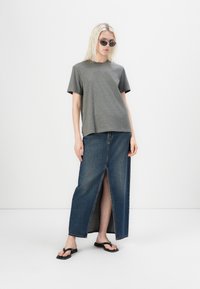 T-shirt gris à manches courtes associé à une jupe maxi en denim taille haute avec une fente avant. Le modèle porte des tongs noires et des lunettes de soleil.