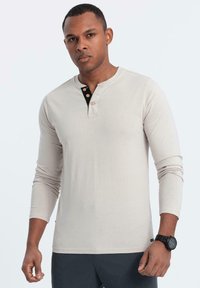 Maglietta henley a maniche lunghe di un beige chiaro, tessuto morbido, con una patta nera dotata di tre bottoni e una vestibilità aderente.