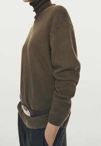 Persona che indossa un maglione verde oliva sopra un dolcevita scuro, con le mani nelle tasche, cintura e pantaloni scuri, su uno sfondo neutro.