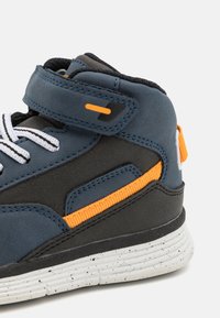 Høy-top sneakers i blått og svart materiale med oransje detaljer. Har hvite snørebånd, borrelås og flekkete hvit gummisåle.