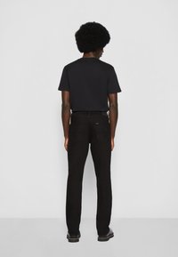 Persona con peinado afro, vistiendo camiseta negra, pantalones vaqueros negros y zapatos negros, de pie frente a un fondo blanco liso.