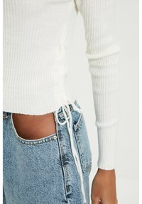 Chemise blanche à manches longues côtelée avec liens sur les côtés, assortie à un jean en denim bleu clair avec bord brut et détails de coutures visibles.