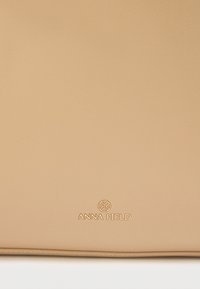 Τσάντα χειρός από μπεζ βινύλ με απαλή υφή, ορθογώνιο σχήμα και ανάγλυφο λογότυπο "ANNA FIELD" στην κάτω δεξιά γωνία.