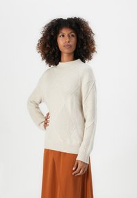 Beige Strickpullover mit hohem Kragen, mit Diamantmustern und gerippten Akzenten. Das Modell trägt einen rostfarbenen Rock.