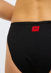 Bas de bikini côtelé noir avec une petite étiquette rouge portant l'inscription « HUGO ». Le design met en avant un look épuré et ajusté avec une texture subtile.