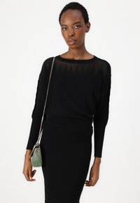 Melna ribota džemperis ar garām piedurknēm, kuram ir caurspīdīga augšdaļa. Modelis tur mazu, teksturētu melnu un zaļu rokassomu.