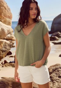 Haut texturé vert olive à manches courtes et en V, associé à un short blanc à revers, sur une plage de sable avec un arrière-plan rocheux.