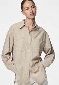 Camicia button-up beige chiaro con strisce bianche, due tasche frontali e un taglio morbido. Realizzata in un tessuto morbido e traspirante.
