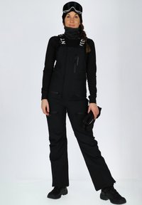 Zwart winteroverall met verstelbare banden, meerdere met rits afgesloten zakken en een zachte textuur. Draagt met zwarte handschoenen en een bril.