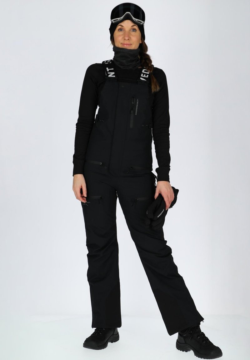 Zwart winteroverall met verstelbare banden, meerdere met rits afgesloten zakken en een zachte textuur. Draagt met zwarte handschoenen en een bril.